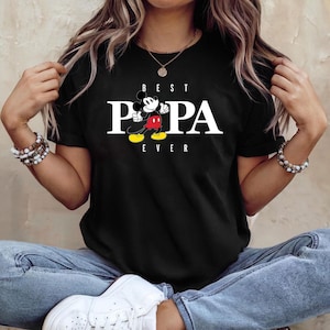 Op de afbeelding: Zwart T-shirt met de tekst "BEST PAPA EVER" in witte blokletters. Een cartoon Mickey Mouse-personage is afgebeeld tussen de woorden "PA" met een rode broek, gele schoenen en witte handschoenen.