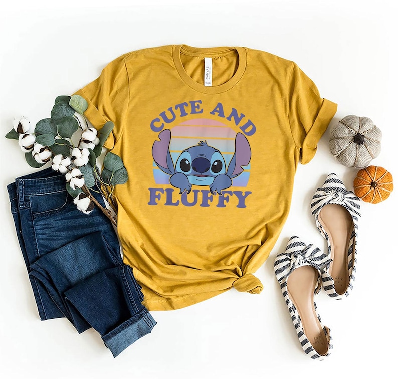 May include: Mustard yellow t-shirt featuring a cartoon character with the text "Cute and Fluffy". The shirt is styled with blue jeans, striped flats, and decorative pumpkins. The shirt is made of a soft material.