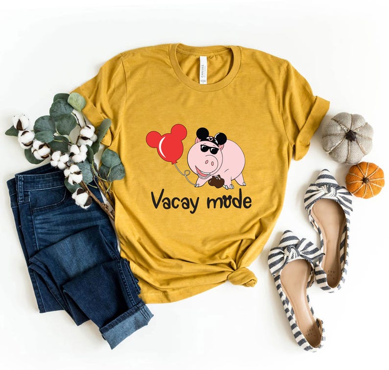 May include: A mustard yellow t-shirt with a cartoon pig wearing Mickey Mouse ears and sunglasses. The pig is holding a red balloon with Mickey Mouse ears. The text "Vacay mode" is printed below the pig.
