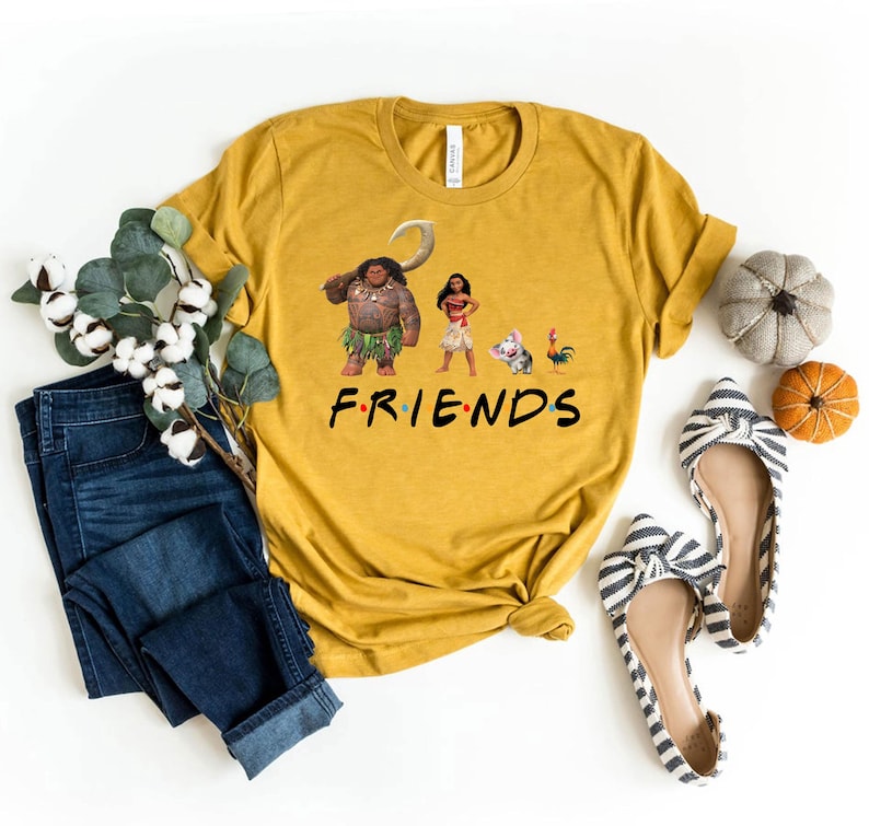 K&ouml;nnte beinhalten: Ein gelbes T-Shirt mit einem Aufdruck von Figuren aus dem Film Vaiana. Der Aufdruck zeigt Maui, Vaiana, das Schwein Pua und einen Hahn. Unter dem Aufdruck steht der Text "FRIENDS".