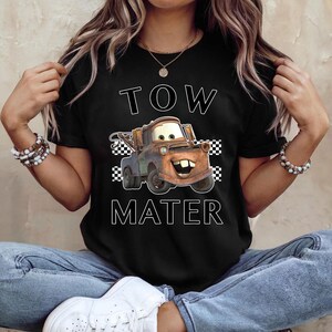 May include: Black t-shirt featuring a cartoon image of Tow Mater from the Cars movie. The design includes the words "TOW MATER" in white block letters, with a checkered flag pattern on either side of the character.