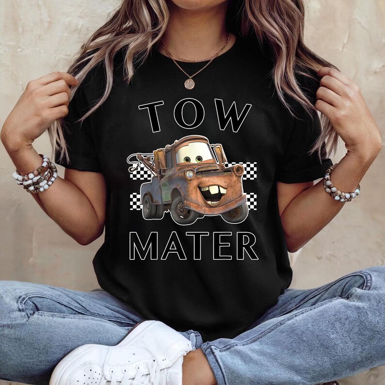 May include: Black t-shirt featuring a cartoon image of Tow Mater from the Cars movie. The design includes the words "TOW MATER" in white block letters, with a checkered flag pattern on either side of the character.