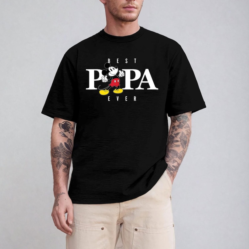 Op de afbeelding: Zwart T-shirt met de witte tekst "BEST POPA EVER" en een Mickey Mouse cartoonafbeelding. Mickey wordt afgebeeld in zijn klassieke pose, met een rode broek, gele schoenen en witte handschoenen.
