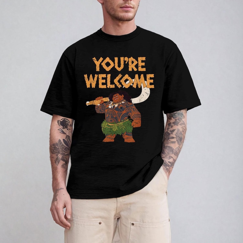 May include: Black t-shirt featuring the phrase "YOU'RE WELCOME" in orange text. The shirt displays a cartoon illustration of a Polynesian figure holding a weapon. The figure is brown with tattoos and a green grass skirt.