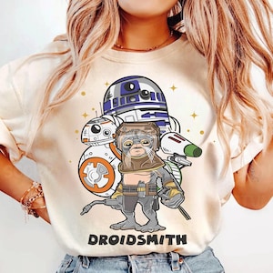 Op de afbeelding: Crèmekleurig grafisch T-shirt met een stripfiguur in een lashelm en uitrusting, met de droids BB-8 en R2-D2 op de achtergrond. De tekst "DROIDSMITH" staat eronder gedrukt. Het ontwerp bevat sterren en een groen en wit element.