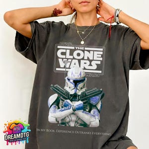 May include: Dark gray t-shirt featuring a graphic from "Star Wars: The Clone Wars." The design includes the show's logo and an illustration of a character in white and blue armor holding two blasters. The text reads, "IN MY BOOK, EXPERIENCE OUTRANKS EVERYTHING."