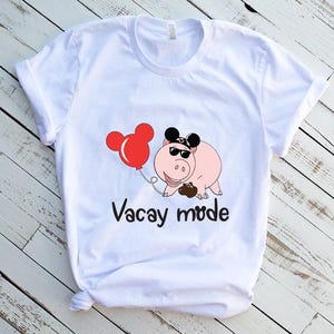 May include: White t-shirt with a cartoon pig wearing sunglasses and a Mickey Mouse hat. The pig is holding a red balloon with Mickey Mouse ears. The text "Vacay mode" is printed below the pig.