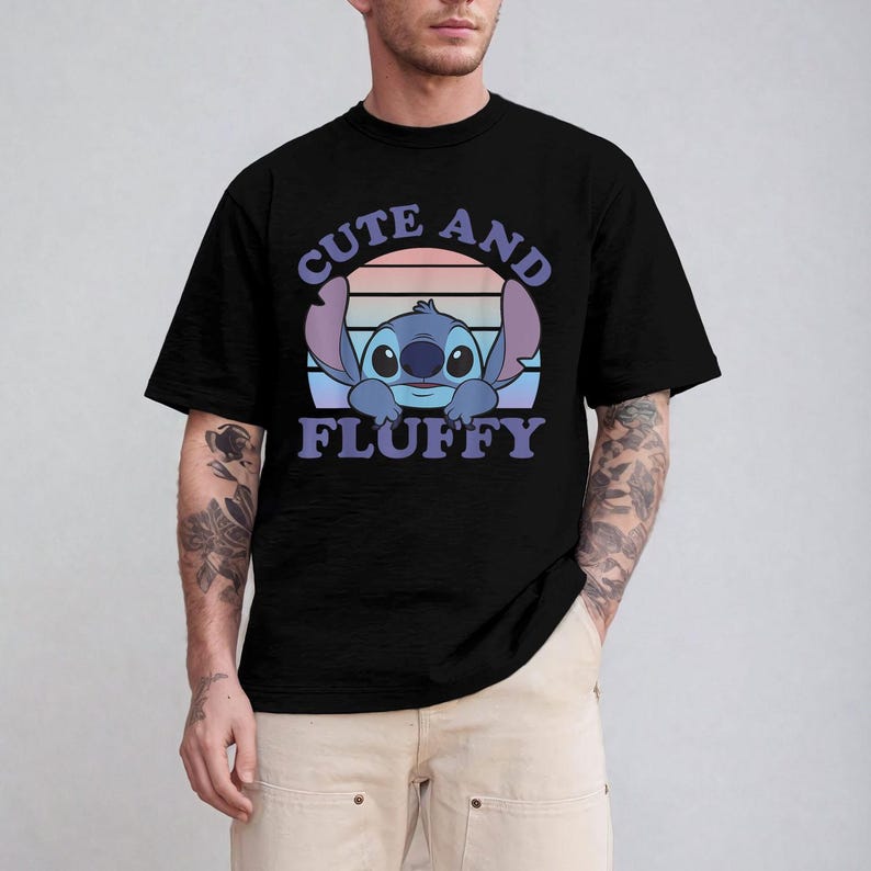 May include: Black t-shirt featuring a cartoon illustration of Stitch with the text "CUTE AND FLUFFY" in purple and blue. The design includes a sunset-like background with pink, orange, and blue hues. The shirt is a crew neck style.