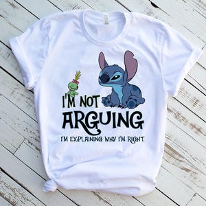 May include: White t-shirt featuring a blue cartoon character and a green alien. The text on the shirt reads "I'M NOT ARGUING I'M EXPLAINING WHY I'M RIGHT". The design is printed on a white t-shirt.