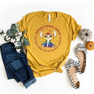 May include: Mustard yellow t-shirt featuring a Stitch graphic and the phrase "OHANA MEANS FAMILY, FAMILY MEANS NOBODY GETS LEFT BEHIND OR FORGOTTEN." The image includes blue jeans, striped flats, and small decorative pumpkins.