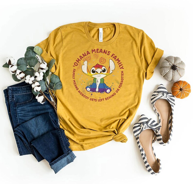 May include: Mustard yellow t-shirt featuring a Stitch graphic and the phrase "OHANA MEANS FAMILY, FAMILY MEANS NOBODY GETS LEFT BEHIND OR FORGOTTEN." The image includes blue jeans, striped flats, and small decorative pumpkins.