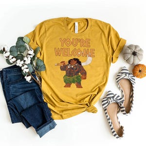 May include: Mustard yellow t-shirt with the text "YOU'RE WELCOME" and a graphic of a cartoon character holding a weapon. The shirt is styled with blue jeans, striped flats, and decorative pumpkins.
