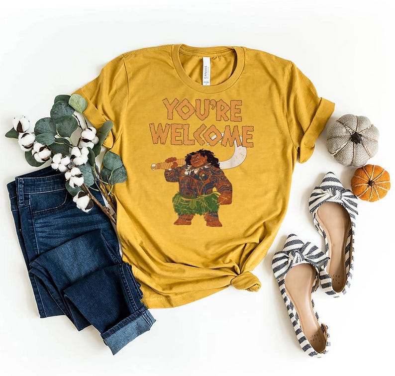 May include: Mustard yellow t-shirt with the text "YOU'RE WELCOME" and a graphic of a cartoon character holding a weapon. The shirt is styled with blue jeans, striped flats, and decorative pumpkins.