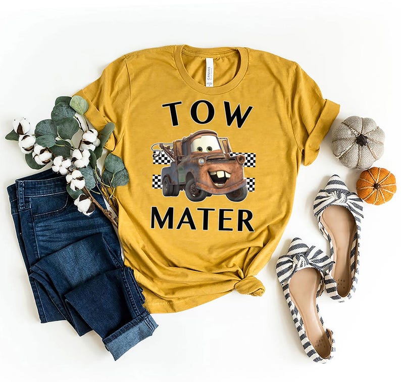 May include: Mustard yellow t-shirt featuring a graphic of Tow Mater from the Cars movie, with the text "TOW MATER" in black. The shirt is styled with blue jeans, striped flats, and decorative pumpkins.
