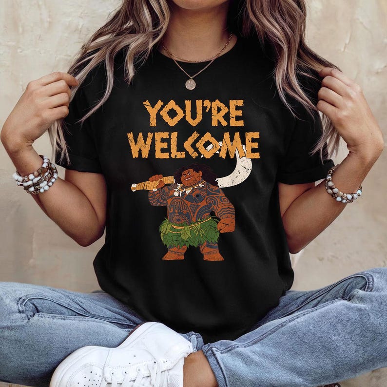May include: Black t-shirt featuring the phrase "YOU'RE WELCOME" in orange, with a cartoon illustration of a Polynesian figure holding a weapon. The figure has tattoos and a grass skirt. The shirt is a casual wear item.