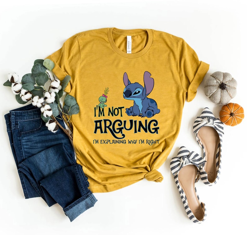May include: Mustard yellow t-shirt featuring a blue cartoon character and the text "I'm not arguing, I'm explaining why I'm right." The shirt is paired with blue jeans and striped flats. Decorative pumpkins and cotton stems are also present.