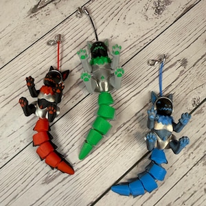 May include: Three colorful, articulated Protogen-style figurines with tails. One is red and black, another is green and silver, and the third is blue and silver. Each has a small clasp for attachment.