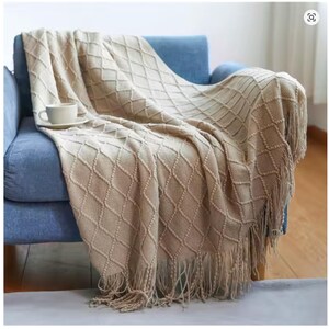 May include: A beige knitted throw blanket with a diamond pattern and fringe detail is draped over a blue armchair. A white teacup and saucer sit on the blanket. The blanket adds a cozy touch to the room.