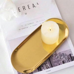 May include: A gold-colored, oval-shaped tray with a lit, white candle. The tray sits on a magazine with the word "EREAL" visible. The candle is cylindrical and has a small flame.