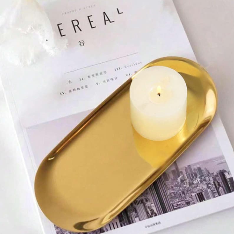 May include: A gold-colored, oval-shaped tray with a lit, white candle. The tray sits on a magazine with the word "EREAL" visible. The candle is cylindrical and has a small flame.