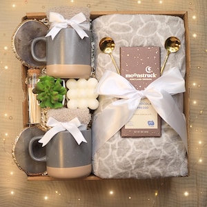 May include: A gift box filled with cozy items, including a gray mug with a brown rim, a white throw blanket, a bar of soap, a small succulent plant, a box of chocolate, and two gold spoons. The box is tied with a white ribbon and has the text "moonstruck portland oregon" on the chocolate bar.