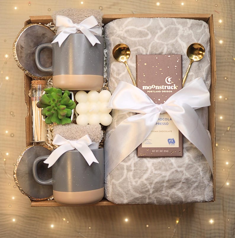May include: A gift box filled with cozy items, including a gray mug with a brown rim, a white throw blanket, a bar of soap, a small succulent plant, a box of chocolate, and two gold spoons. The box is tied with a white ribbon and has the text "moonstruck portland oregon" on the chocolate bar.