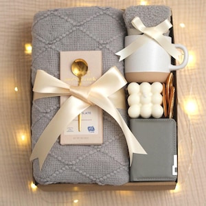 May include: A gift box with a gray blanket, a white and tan mug, a small gray wallet, a gold spoon, a bar of chocolate, a small gray towel, a white candle, and dried orange slices. All items are tied with a cream-colored ribbon.