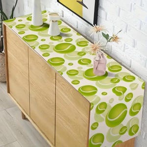 May include: A white table runner with a green tennis ball pattern. The runner is on a wooden table with a vase of flowers.