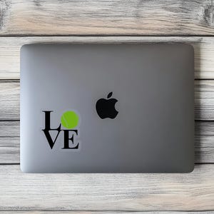 May include: A laptop computer with a black and white sticker that says "LOVE" with a tennis ball in the "O". The sticker is on the silver laptop.