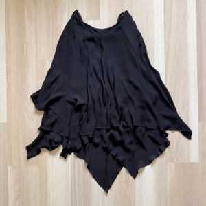 May include: A black skirt featuring a layered, asymmetrical hemline with multiple flowing panels. The garment is constructed from a lightweight, semi-sheer fabric that creates soft folds and drapes throughout the design, ending in pointed edges.