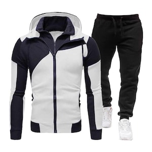 Può includere: Un completo tuta sportiva composto da una felpa con cappuccio e zip e pantaloni abbinati. La felpa è bianca con inserti blu navy, e i pantaloni sono neri. L'outfit è completato da scarpe da ginnastica bianche.