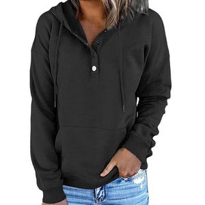 Könnte beinhalten: Ein schwarzer Kapuzenpullover mit Knopfleiste und Vordertasche. Das langärmlige Oberteil hat eine Kapuze mit Kordelzug und gerippte Bündchen. Die Person trägt eine blaue Jeans.