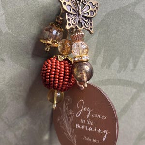 May include: A decorative keychain with a butterfly charm, beads, and a brown tag. The tag has the text "Joy comes in the morning" and "Psalm 30:5" in white script. The keychain features a mix of gold, brown, and red elements.