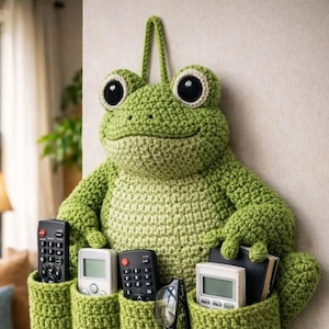 May include: A whimsical, handmade crochet frog wall organizer in shades of green. The frog has large, expressive eyes and holds four pockets, perfect for storing remotes, glasses, and small items. A fun and functional home decor piece.