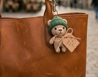May include: A tan leather handbag with a small crocheted bear keychain attached to the handle. The bear wears a green knit hat and has a small brown paper tag tied around its neck with twine, featuring the text Made with love for you.