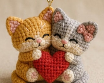 May include: A handmade crochet keychain featuring two smiling cat figures, one orange and one gray, holding a red heart. The cats have pink accents on their ears and a gold-colored keyring.