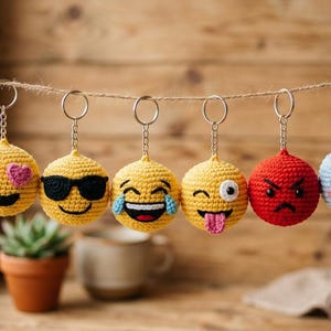 Puede incluir: Una colección de llaveros emoji hechos a mano con ganchillo. Los llaveros presentan varias expresiones, incluyendo caras sonrientes, corazones, gafas de sol y una lengua fuera. Los llaveros son amarillos, rojos y azules, y están colgados de una cuerda.