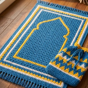 May include: A blue crochet prayer rug with a yellow and white border and a matching drawstring bag. The rug has a central arched design in yellow. The rug and bag are on a wooden floor.