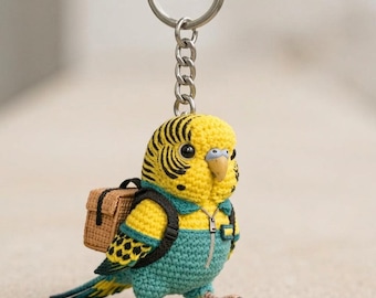 May include: A handmade crochet keychain featuring a yellow and teal parakeet wearing a small backpack. The parakeet has black stripes on its head and is attached to a silver keyring. The bird is wearing a teal jumpsuit with a zipper.