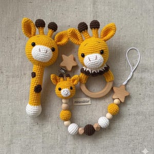 May include: A set of handmade crochet baby toys in the shape of giraffes. The set includes a rattle, a teether, and a pacifier clip. The toys are yellow with brown spots and accents, and white faces. The pacifier clip has wooden beads and a star.