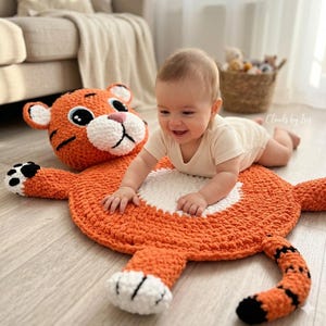 Peut inclure: Tapis en crochet orange et blanc en forme de tigre, avec un bébé souriant. Le tapis représente une tête de tigre, des pattes et une queue rayée. Le bébé porte un body clair et est allongé sur le tapis.