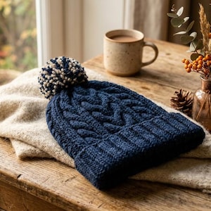 Può includere: Un berretto blu navy a trecce con un pompon color crema è appoggiato su una sciarpa color crema. Una tazza di caffè, un piccolo vaso di fiori secchi e pigne sono su un tavolo di legno vicino a una finestra.