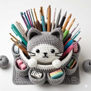 Può includere: Un organizer a forma di gatto all'uncinetto grigio e bianco, pieno di penne, uncinetti e materiali per l'artigianato. L'organizer ha piccole tasche per dadi, post-it e gomme. Due gomitoli di filato grigio su entrambi i lati.