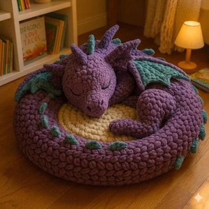 Könnte beinhalten: Ein großer, gehäkelter lila Drache, der sich auf einer runden, cremefarbenen Matte zusammenrollt. Der Drache hat türkisfarbene Flügel und Akzente, die Augen sind geschlossen. Der Drache ist handgefertigt.