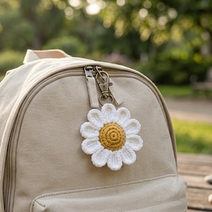 Op de afbeelding: Een beige rugzak met een sleutelhanger in de vorm van een madeliefje. Het madeliefje is wit met een gele kern. De rugzak staat op een houten oppervlak met een vage achtergrond van bomen en zonlicht.
