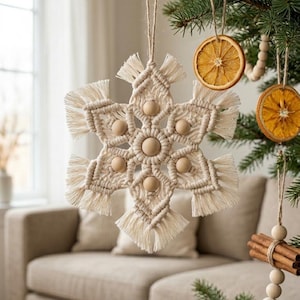 May include: A handmade macrame snowflake Christmas ornament with wooden beads. The off-white snowflake is hanging on a Christmas tree with other natural ornaments, including dried orange slices and cinnamon sticks. The background includes a couch and a window.
