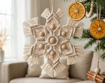 May include: A handmade macrame snowflake Christmas ornament with wooden beads. The off-white snowflake is hanging on a Christmas tree with other natural ornaments, including dried orange slices and cinnamon sticks. The background includes a couch and a window.