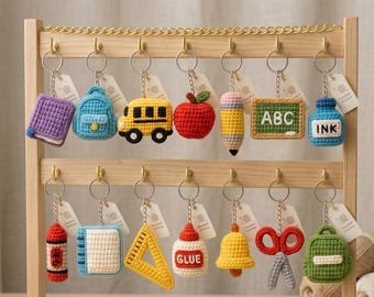 May include: A wooden display stand holds a collection of crocheted keychains. The keychains feature school-themed designs, including a book, backpack, school bus, apple, pencil, chalkboard with "ABC", and ink bottle. Other keychains include a crayon, notebook, ruler, glue, bell, scissors, and backpack.
