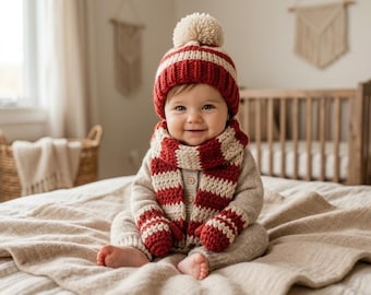 May include: A baby wearing a red and white striped knit hat, scarf, and mittens, with a matching pom-pom. The baby is also wearing a beige sweater and is sitting on a beige blanket. A crib and window are in the background.