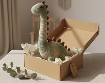 May include: A crocheted dinosaur toy in a cardboard box. The dinosaur is primarily light green with brown accents along its back. The box is open, revealing the toy nestled in white paper. A small tag hangs from the box.